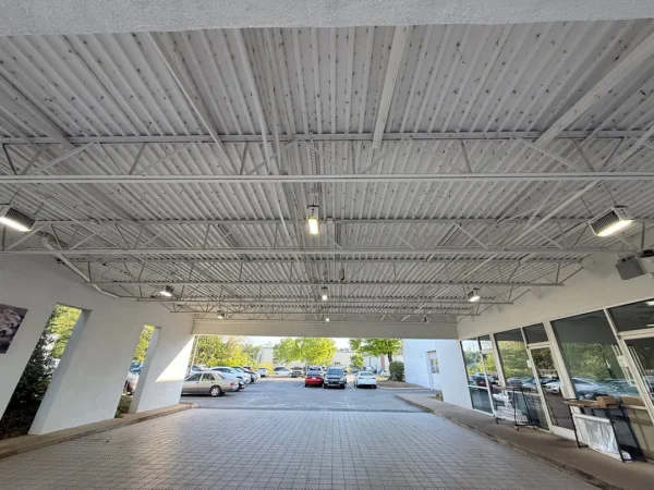 Covered driveway with exposed metal beams and hanging lights leading to a parking lot with cars outside the building's glass entrance area.