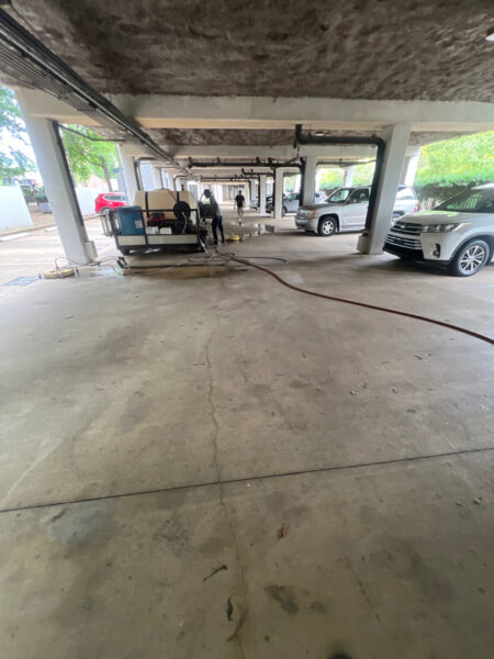 Maintenance workers in a concrete parking garage with hoses and a mobile service cart near parked SUVs, performing a cleanup or service task.