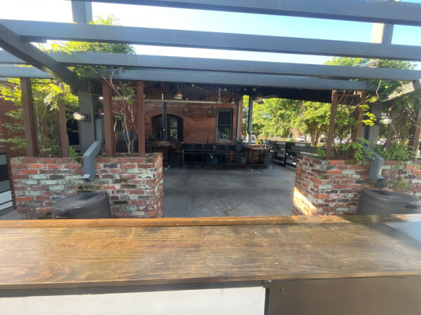 Open-air brick patio with a wooden bar counter in the foreground and a seated dining area under a metal-framed pergola.