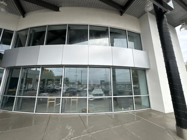 Curved glass storefront of a modern building with metal panels and a parking lot reflection in the windows