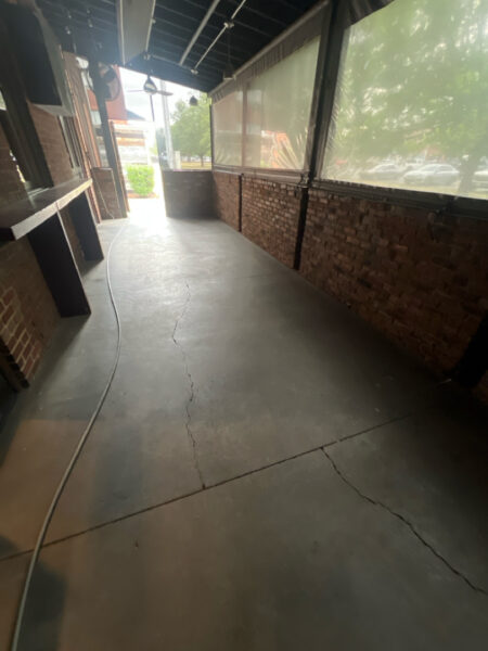 Covered outdoor walkway with brick walls, a coiled hose on the concrete floor, and a view of greenery at the far end.