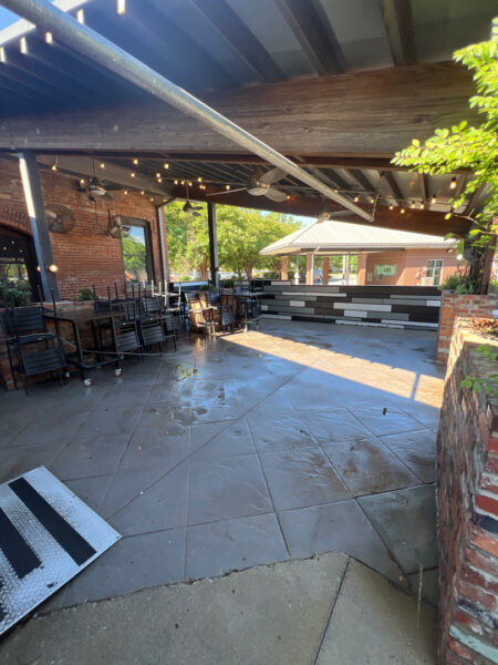 Covered outdoor dining patio with brick walls, string lights, and stacked black tables and chairs.