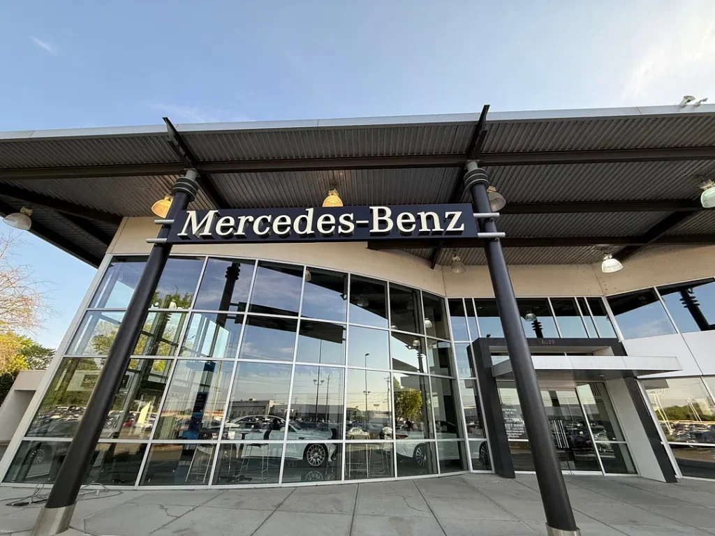 Mercedes-Benz dealership with a glass-front showroom, dark diagonal columns, and cars visible inside the windows.