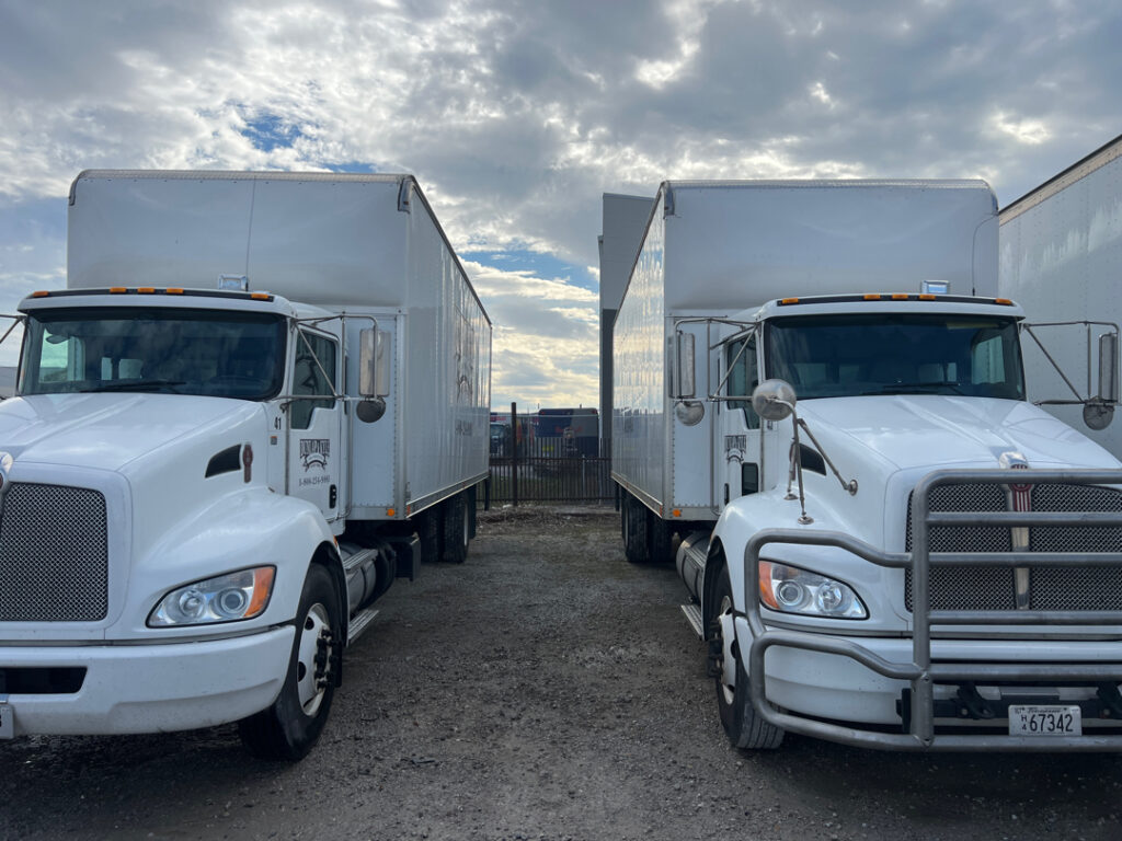 Two white semi-trailers parked side by side in a gravel lot under a cloudy sky, front ends toward the camera.