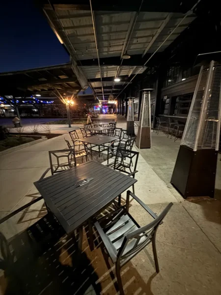 Nighttime outdoor dining area with empty metal tables and chairs under a covered walkway, with tall patio heaters nearby.