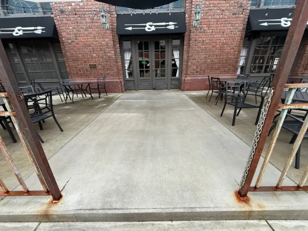 Outdoor cafe seating on a concrete patio in front of a brick building with black awnings and glass doors.