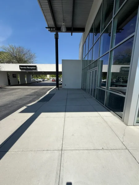 Sidewalk along a glass-front building with a tall black column and overhang; a distant 'Service Reception' sign under a parking canopy.