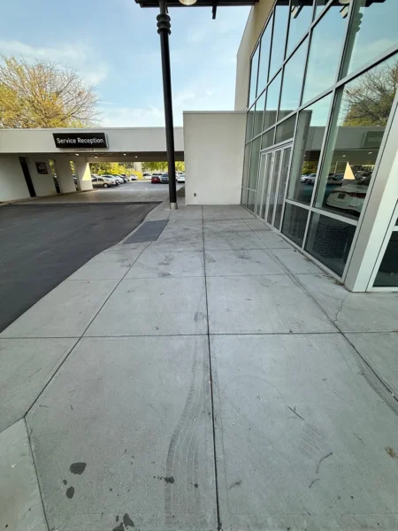Wide concrete sidewalk in front of a modern glass-fronted building with a sign reading 'Service Reception' and parked cars beyond the driveway.