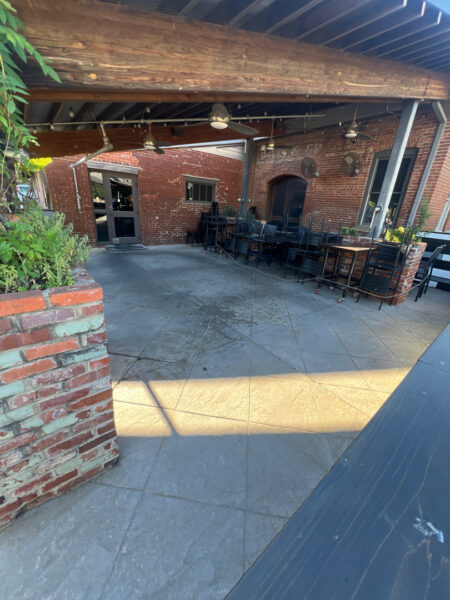 Outdoor brick patio with stacked chairs along a long table under a wooden beam and ceiling fans overhead.