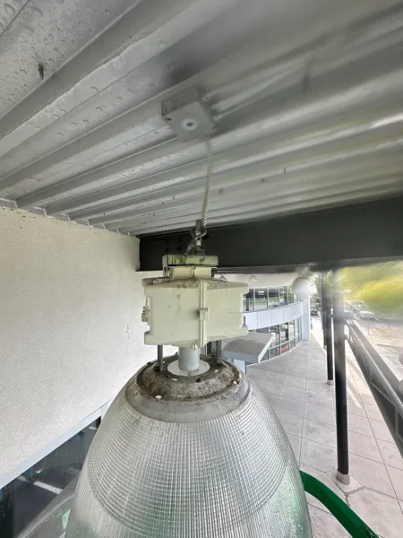 Outdoor industrial floodlight with a weatherproof glass dome mounted under a metal ceiling, connected to a white electrical transformer box above the lamp.