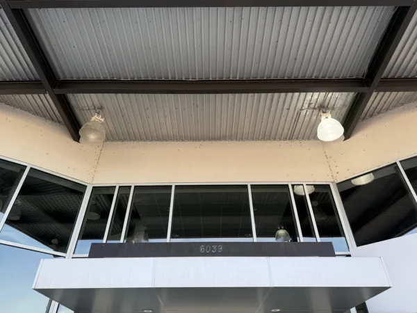 Exterior view of a modern building entrance with a metal overhang, large glass storefront windows, and two hanging industrial lights; address 6039 visible on a dark sign.