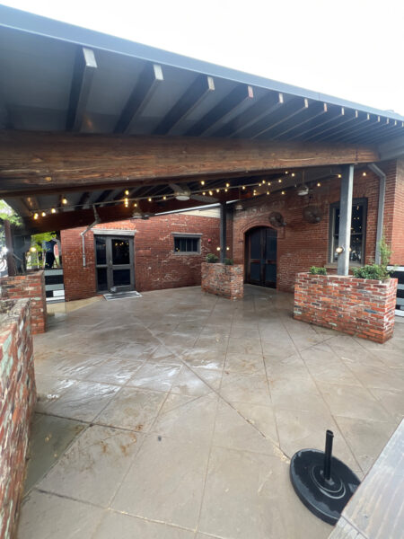 Brick-walled outdoor patio with a covered roof, string lights, and planters along brick walls near black doors.