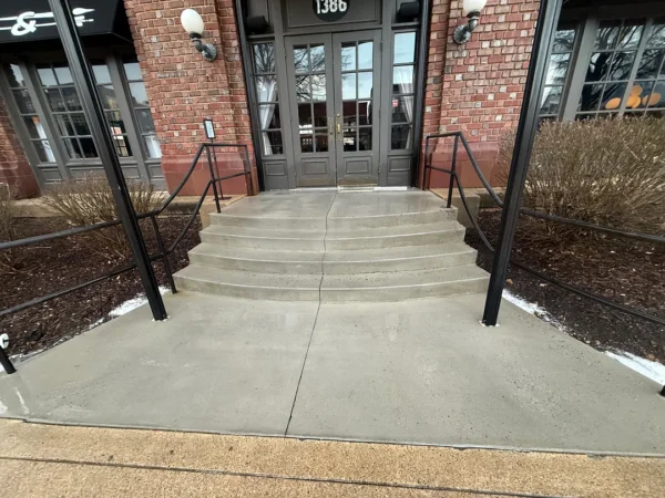 Brick building entrance with curved concrete steps, black metal handrails, and double doors at the top.