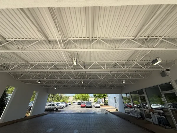 Interior of a covered drive-through area with an exposed white metal truss ceiling and hanging lights; cars parked outside beyond the opening.