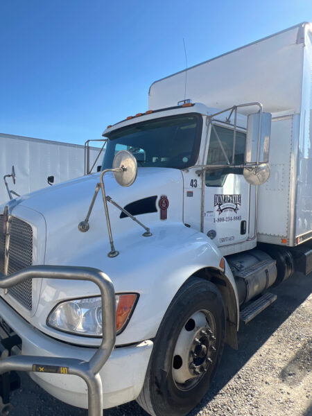 White Kenworth semi-truck tractor with a box trailer, chrome mirrors, and a front bumper guard parked on a gravel lot.