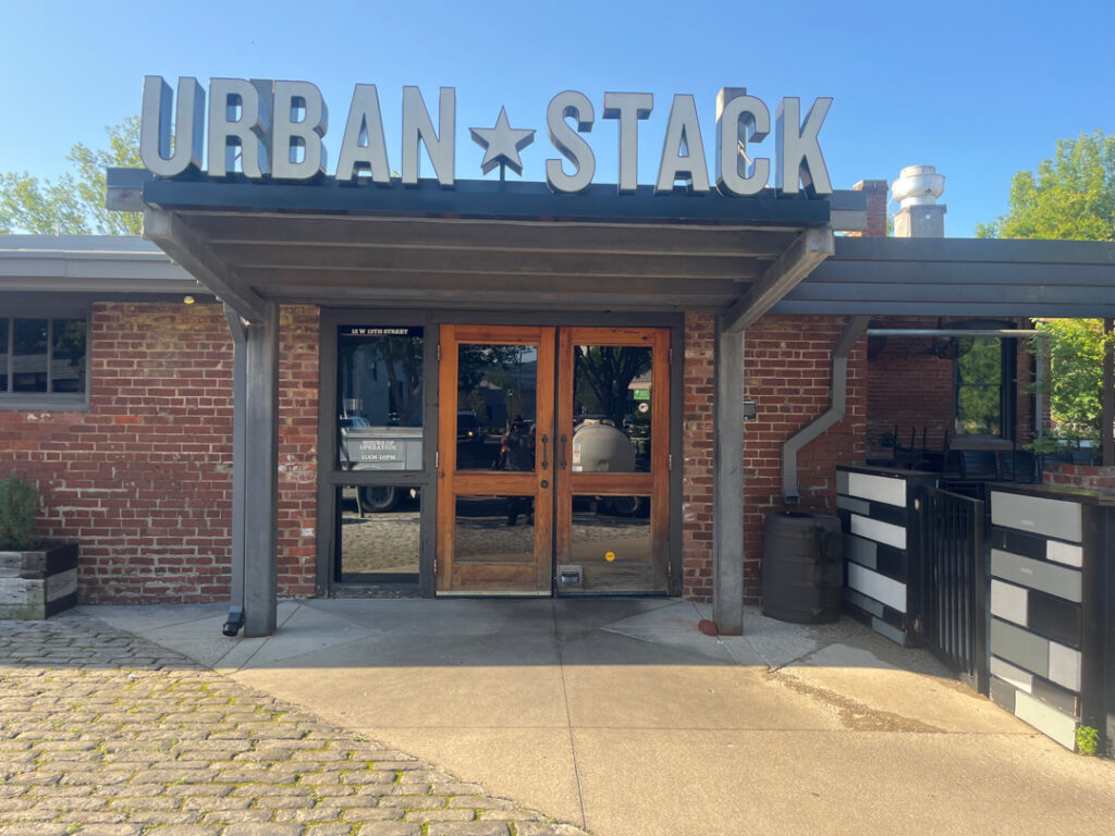 Front entrance of Urban Stack restaurant with brick exterior and double wooden doors under a metal canopy, bright blue sky above.