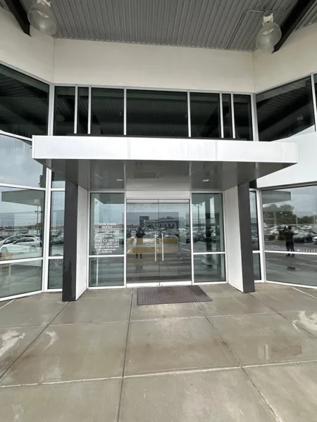 Glass storefront entrance with double doors under a silver canopy; showroom windows reflect parked cars outside.