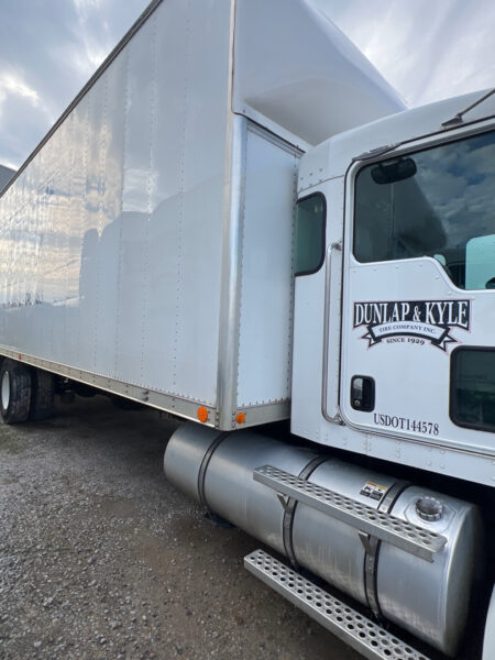 White semi-truck with a large white trailer attached, featuring a Dunlap & Kyle logo on the cab door and a metallic fuel tank with steps in the foreground on a gravel surface