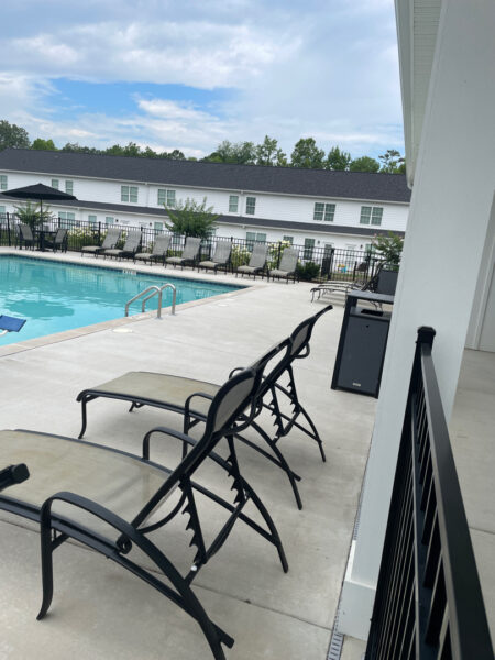 Poolside patio with several black lounge chairs on a concrete deck, blue pool to the left, and a white building with multiple windows in the background.