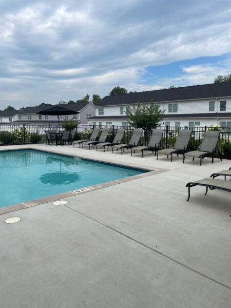 Outdoor apartment pool area with a row of lounge chairs along a fence and a table with a black umbrella.