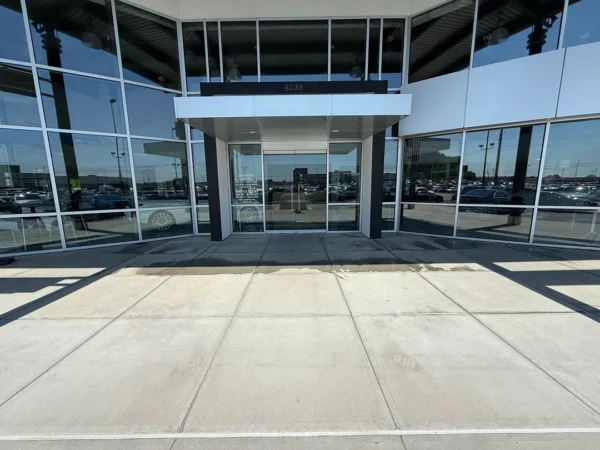Front entrance of a glass-walled office building with a white canopy over double glass doors, reflecting a parking lot in the windows (address 6039 visible).