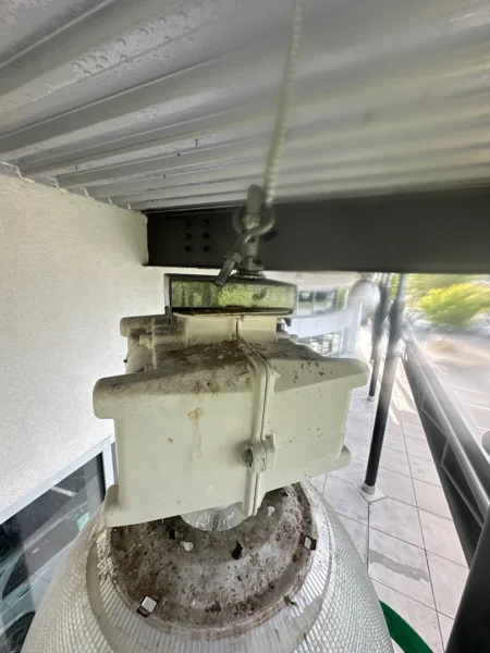 Outdoor ceiling-mounted light fixture with a dusty, rusted housing and weathered base under an overhang.