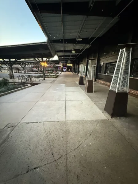Wide sidewalk under a covered plaza with outdoor seating to the left and tall patio heaters along the right storefronts.