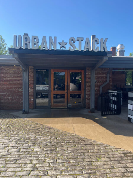 Urban Stack storefront entrance with large metal sign and star, brick exterior under a blue sky, double wooden doors.