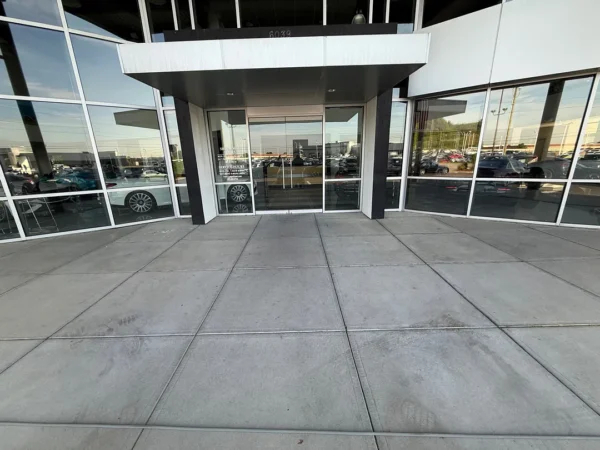 Wide view of a car dealership entrance with glass doors, flanked by large windows reflecting parked cars; a wide concrete sidewalk stretches to the foreground.