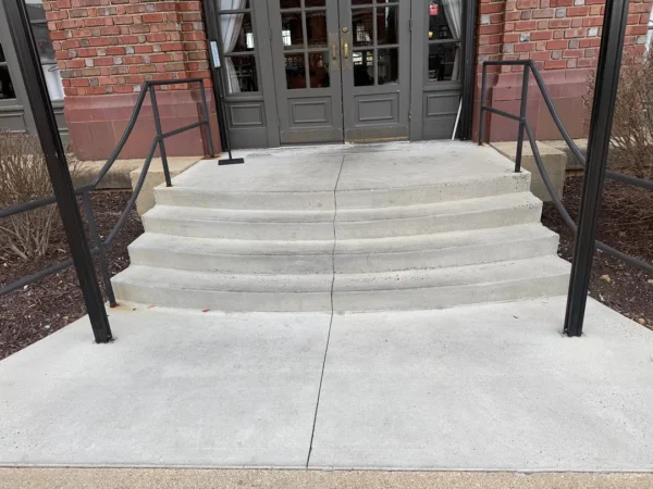 Concrete steps leading to glass doors with black metal handrails on both sides, featuring a visible center crack in the middle of the stairs.