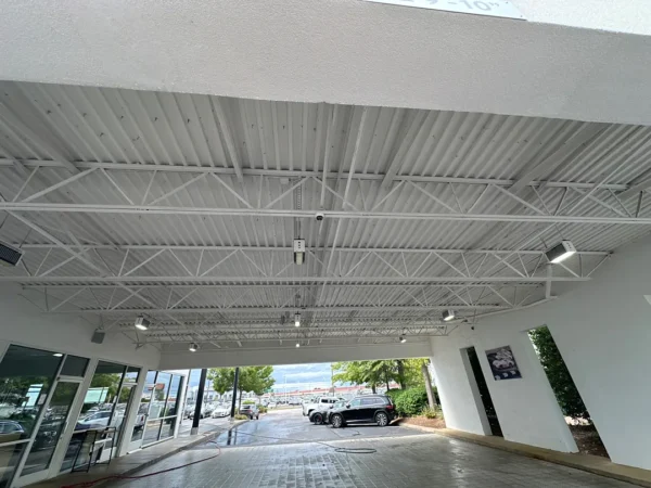 Covered outdoor entrance with white metal trusses and ceiling lights; glass storefront on the left and parked cars beyond, with trees and a poster on the wall
