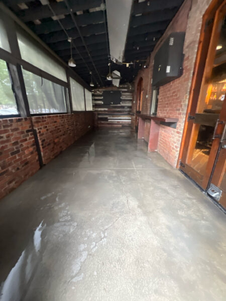 Empty industrial-style hallway with brick walls, concrete floor, and exposed ceiling; windows along the left and a door on the right.