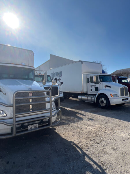Front view of a white semi-truck with a heavy grille guard in a gravel truck yard on a sunny day.