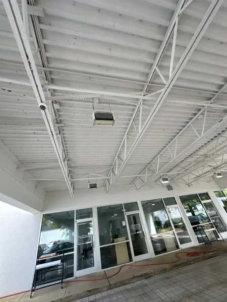 Exterior storefront under a white metal roof with exposed trusses and a glass-entry door; a red hose lies along the sidewalk outside.