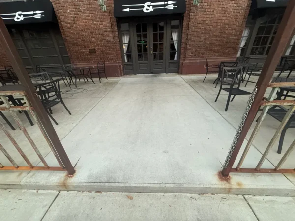 Brick storefront with black doors, outdoor seating, and rusty metal railings on a concrete patio outside a cafe.
