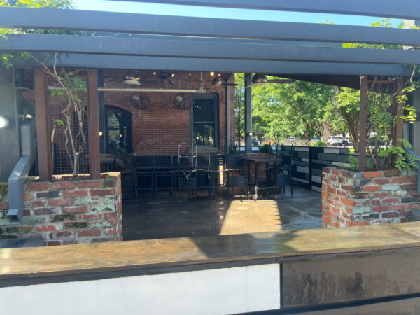Outdoor cafe patio with brick walls, black metal chairs and tables under a wooden pergola on a sunny day.