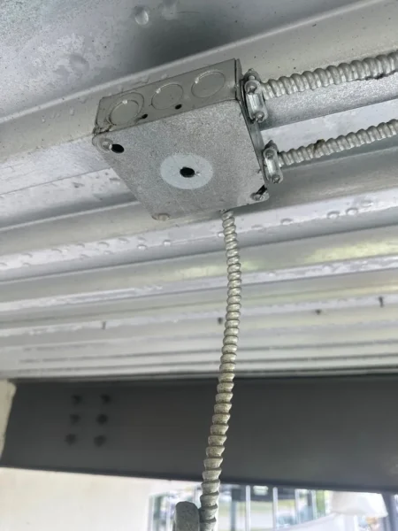 Metal electrical junction box mounted to a galvanized beam with flexible conduit running alongside it; small circular knockouts visible on the box.