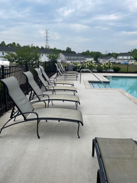 Row of empty gray lounge chairs along a concrete pool deck beside a fenced swimming pool on a cloudy day.