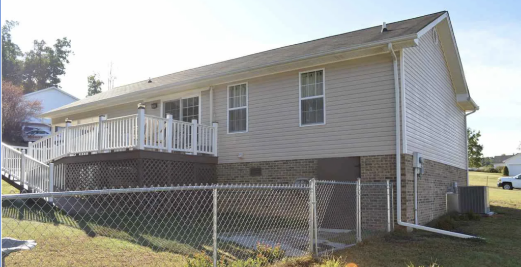 Two-story beige house with a raised white deck and railing, brick foundation, and a chain-link fence in front yard.
