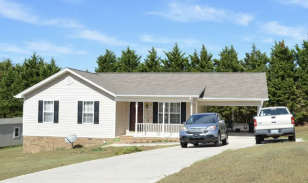 Front view of a single-story white house with a small porch, two windows with shutters, and a concrete driveway with a silver SUV and a white pickup parked outside. Behind are tall evergreen trees.