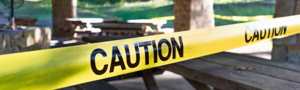 Yellow caution tape blocks off a pavilion with wooden picnic tables in the background restricting access to the area
