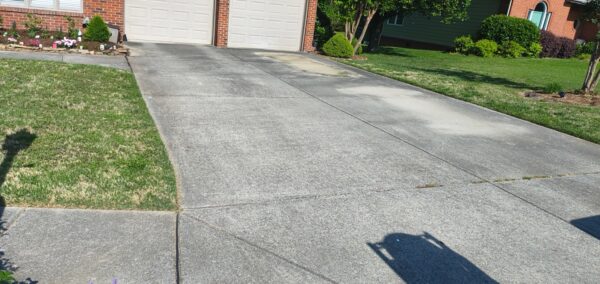 Concrete driveway leading to a two-car garage, with green lawns and shrubs on the sides; photographer's shadow in the lower right.