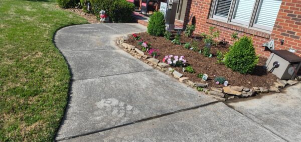 Curved concrete walkway beside a brick house, bordered by a mulched flower bed and rocks, with green lawn on the left.