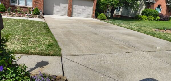 Residential driveway leading to a two-car garage with brick house, manicured lawn, and flowering border on the left; a sidewalk and a sharp shadow from a street sign are visible.