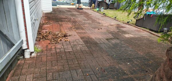 Red brick patio beside a house with a pile of dried leaves near the wall and patio furniture in the background near a pool area.