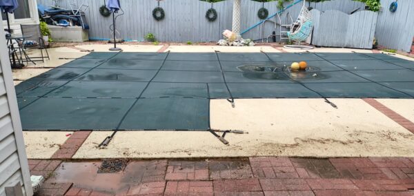 Backyard swimming pool with a green safety cover secured by straps and puddles on the surface.