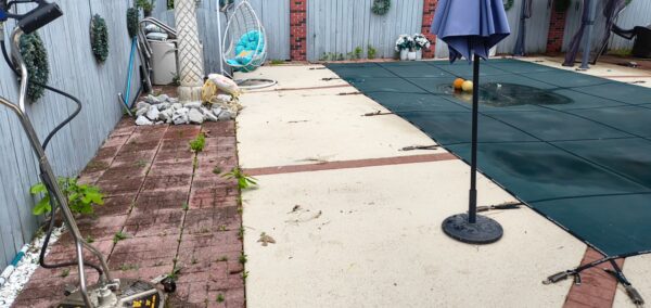 Backyard scene with a covered pool along a beige concrete patio, a dark pool cover, and a blue wooden fence with plants and tools nearby.