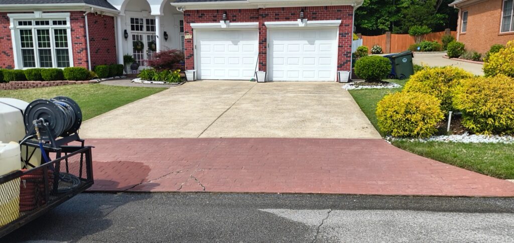 Front of a red-brick house with two white garage doors, concrete and brick driveways, and manicured landscaping on a sunny day.