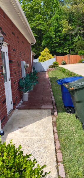 Brick house side yard with a narrow brick walkway, potted plant, and row of trash bins along the lawn.