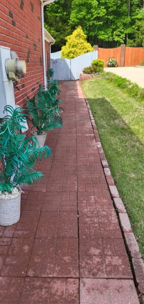 Red brick walkway running beside a brick house, with potted green plants along the wall and a gray wooden fence at the end; a grassy lawn to the right.